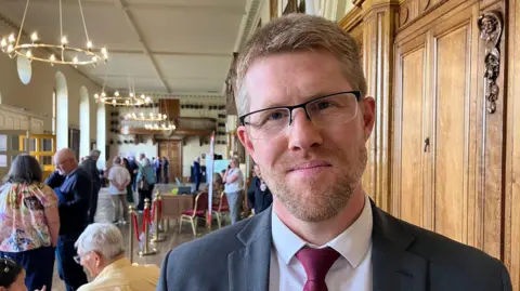 A man with short light brown hair and beard and glasses is wearing a white shirt and wine-coloured tie under a grey suit jacket.