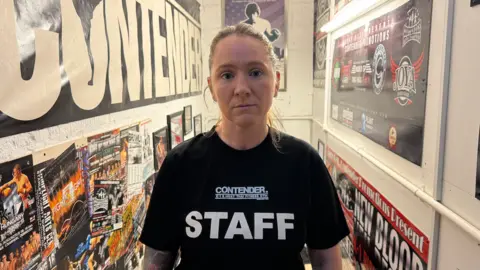 Vicky Caven, wearing a black T-shirt saying "staff" and with the gym's name on it, standing in a hallway at the gym's entrance that is mostly white with gym-themed posters on the walls, saying things like "contender" and "new blood".