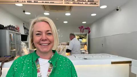 Joanne Martin is smiling brightly at the camera. She has short blonde hair and is wearing a green shirt and a lanyard which reads: "Be Kind." The counter of the fish and chip shop behind her is white and grey. There is a worker in the background, working at one of the stands.