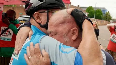 Steve Huntley/BBC Mark Monaghan is on the left hugging one of his friends who just finished the bike ride. Mr Monaghan is wearing a navy blue T-shirt, and the other biker is wearing a light blue T-shirt with a black helmet.
