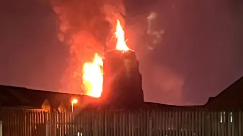 An image of the Dongeall Road bonfire, in the middle distance, beginning to catch light. Flames can be seen rising from the top of it. A metal fence is in the foreground