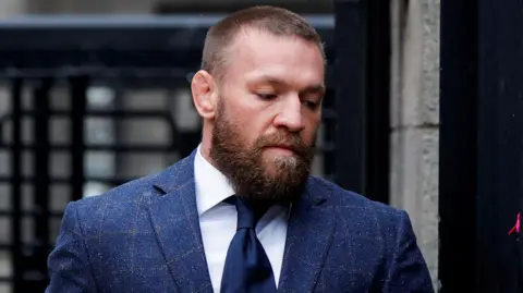 Conor McGregor, who has short, light brown hair and a thick brown beard and wears a blue-checked suit jacket over a white shirt and a navy tie.