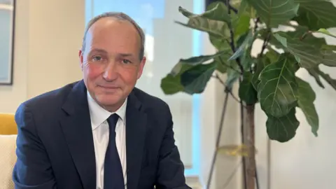 Lyndon is in a dark suit and tie sits on a yellow chair in a modern office, with a large leafy plant and framed artwork behind them.