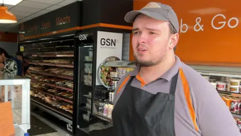 Corin Allen is standing in front of food shelves selling meat and pre-made sandwiches and drinks. He is wearing a black apron with a grey hat and shirt with orange detailing. 