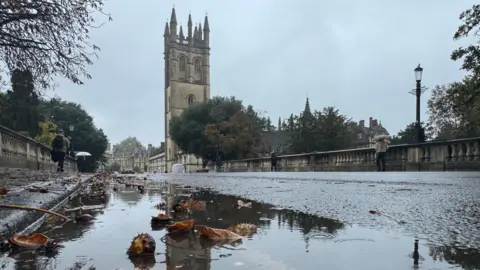 EstherJ Oxford's High Street with a tower in the background. The curb has a puddle running along it, with leaves, branches and conkers in it.