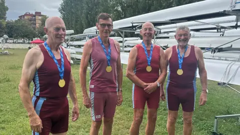 Four men wearing red rowing vest and short with gold medals. They are stood near boats on grass.