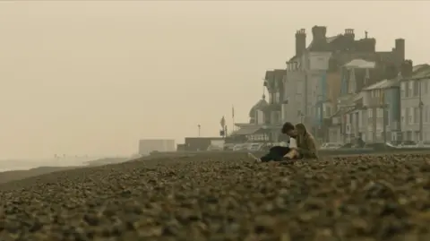 Max Morgan A young man and a young woman sitting on a shingle beach at a hazy time of day. There are seafront buildings behind them.