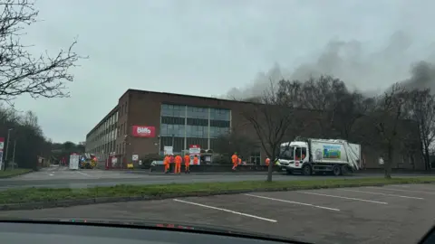 Biffa's three-storey brown-brick building, with Biffa written in white on a red sign on its front wall. Smoke can be seen billowing from behind the building and about seven workers in bright orange fluorescent jackets and trousers standing in a car park in front of the building. A fire engine can be seen parked to the side.