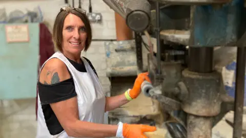 A woman with bobbed hair, with glasses perched on top, wears a white overall. She has orange gloves on, and is holding a handle on a piece of machinery