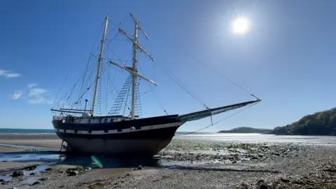 A side-on view of a traditional tall ship sitting upright on a sand and rocky beach with the tide out, run aground. It is a gloriously sunny day with blue skies.