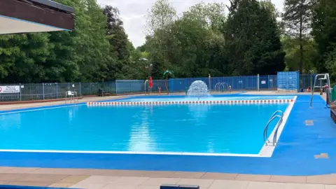 A view of an outdoors swimming pool with a small fountain at one end. Trees surround the perimeter of the pool. 