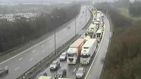 A blurry traffic camera shows lorries and cars queueing as far as the eye can see. The road looks slightly damp with trees to the right side and an empty carriageway on the left. 