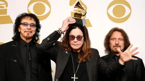 Bandmates Tony Iommi, Ozzy Osbourne and Geezer Butler pose in front of yellow and white Grammy logos while holding a grammy award for Best Metal Performance at the 2014 Grammy awards