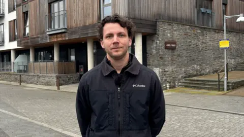 A man with dark hair in a black coat is in the centre of the frame. He has a stern look on his face. Behind him is a block of properties with wooden frontage.