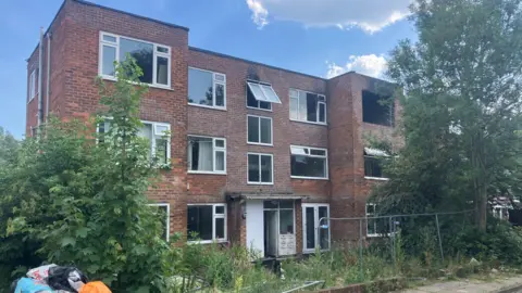 The abandoned apartment block on Baguley Crescent. It is a three-storey red brick building with a flat roof. The central section is set in slightly from the two wings either side. The side elevations have large white framed windows, while replicated either side of the entrance way in the central section. Above that entrance are smaller, double paned windows, the top one of which is open. The image shows burnt-out flats with obvious fire damage on the far right hand section, broken windows and insecure fencing. Plants and trees have overgrown in front of the building and pile of rubbish just comes into shot in the bottom left corner.