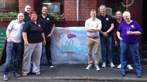 Village Bakers Eight people stood outside a red brick building next to a banner that says Village bakers in blue writing on a white background.