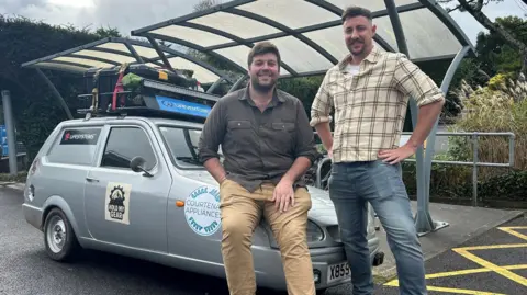 BBC Two men in front of a Reliant Robin vehicle. The man on the left is wearing a chequered shirt and blue jeans, he is holding his hands on his hips. The man on the left is sitting on the bonnet of the vehicle. He is wearing beige chinos and a brown shirt. The car is silver and has a number of stickers on its chassis. There is a roof rack on the top of the car.