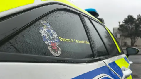 BBC A close up view of the side of a police car. It shows a window and the Devon and Cornwall Police coat of arms. 
