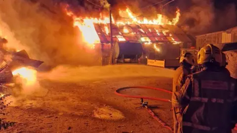 The backs of three firefighters can be seen in the foreground and a large buildng is on fire in the background.