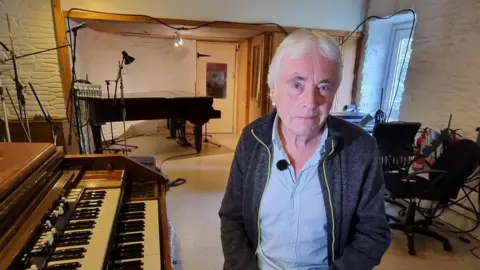 Man sat next to an organ with a piano, music stands and chairs in the background