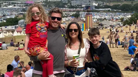 Family handout Kelly, her husband and children at Glastonbury Festival. Her husband is second from left and wearing a grey t-shirt and shades, holding their young daughter in his arms, she is wearing a red Glastonbury t shirt and red leggings. Kelly is wearing a white t shirt, sunglasses and holding a cup of drink and their son is wearing a dark top. The whole family is smiling at the photographer with some bright rainbow tents in the background.