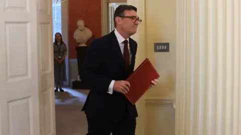 PM Andy Burnham wearing a dark suit walks into a room, holding a red folder.