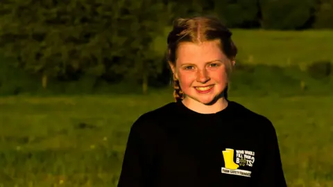 Costas Andreou Erin Thompson stands in a field. She has blond hair tied back in two plaits, blue eyes and freckles. She is wearing a black t-shirt with a logo of two yellow wellies. She is smiling at the camera