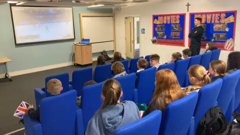 Pupils sitting in blue cinema-style seats watch footage from the Winter Olympics, which is shown on a large projector screen. 