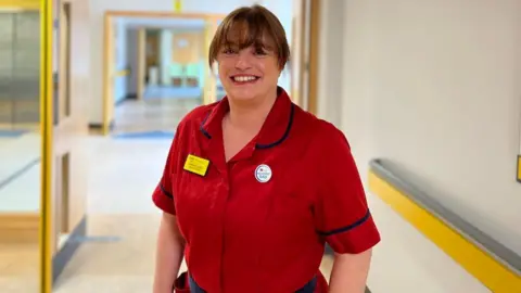 Emma Cleator, a woman with brown hair in a bun and a fringe wears a red Matron uniform and smiles brighlty in a hospital corridor.