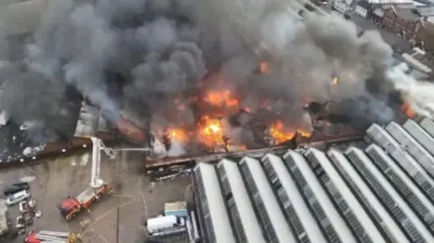 West Midlands Fire Service A photo of an industrial site on fire with a fire engine shooting water onto the fire. There is visible orange flames and thick black and grey smoke coming out of the top of the building. 