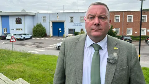 Geoff Willetts of the Prison Officers Association. He is wearing a green suit with some badges on the lapel. He is standing on a hill with Swinfen Hall prison behind him.