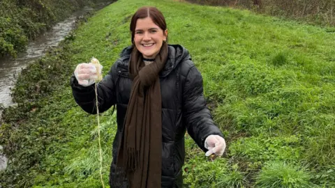 Janet Sturgess A woman, standing by water, smiling, with her arms out, holding onto an item on string. She has long dark hair tied back, wearing a black cat, brown scarf and gloves. A grass bank is behind her. 