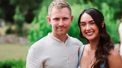 Joe and Jessie smiling into the camera. Joe is standing on the left and has blonde, short hair with stubble around his mouth. He is wearing a cream shirt and is smiling with his mouth closed. Jessie is on the right and has long, dark hair which has been curled. She is smiling with her teeth and has red lipstick on. She is wearing two gold necklaces and a blue square-neck top with wide straps. They are in a garden, but the background is out of focus.