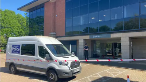 The Solent Centre is a complex of glass-fronted office buildings with brick pillars supporting ground-floor covered walkways. It has police tape around it and a police van outside blocking the entrance.