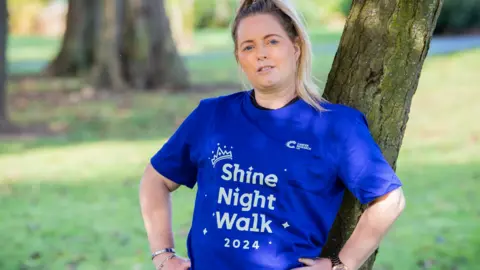 Cancer Research UK Stacey, wearing a royal blue Cancer Research UK t-shirt, stands outside with her back to a tree 