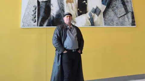 Bradford Council Artist Marcus Levine stands in front of his artwork and a yellow wall. He is wearing a black leather coat and tinted glasses