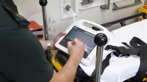 NHS A paramedic in an olive green uniform looks down at a screen as they make records inside an ambulance. Only their shoulders and arms and hands are visible. They are standing over an empty bed with white sheets and black straps over it.