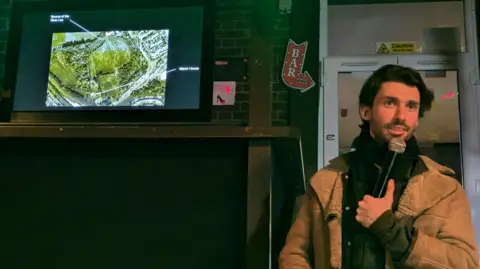 Lucy Wood Nature Calling Matthew Rosier, holding a microphone, standing by a television screen that shows an ancient site. He has short dark hair, is wearing a dark top and jumper and camel coloured coat. He is standing to the right of the screen, in front of a door. He is talking. 