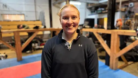 A woman with red hair smiles and looks into the camera. She is standing in the wooden ring where the fights take place.