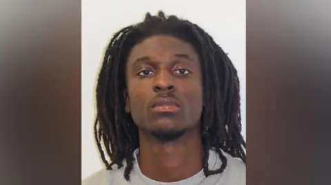 Met Police Custody photograph showing a man with long dreadlocks wearing a grey top against a plain background.