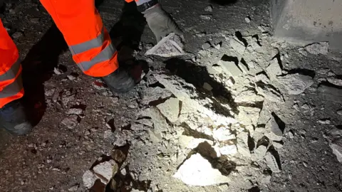 Pieces of grey concrete in a pile on the floor full of rubble. One man's feet can be seen in the picture, with high vis trousers on, and a hand can be seen picking up one of the fragments. The fragment is illuminated by torchlight.
