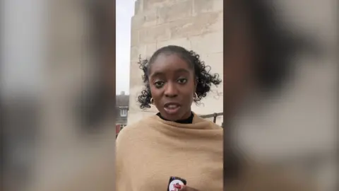 A woman with curly black hair is wear a camel-coloured wrap. She is speaking into the camera and holding a button mic.
