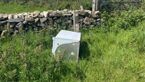 A white fridge freezer is on its side in a countryside setting with long grass, a stone wall and wooden post
