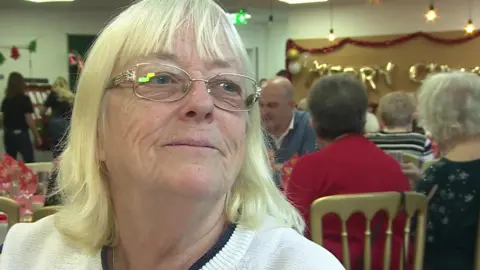 Chris is sitting at a table surrounded by other pensioners. She's wearing a white jumper with navy stripes and she has glasses on.