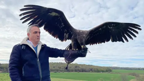 A man in a dark blue zip up coat, and with short white hair, has a falcon on his outstretched left arm with its huge brown wings outstretched