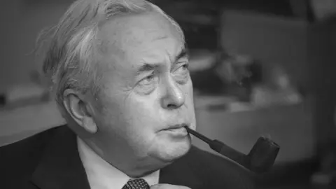 Harold Wilson looks away from the camera, holding a pipe in his mouth. He is wearing a suit.