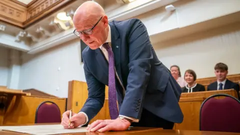 PA Media John Swinney signs a document while hunched over a wooden table, with smiling people looking on 