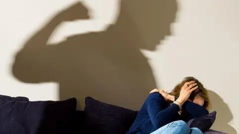 PA Media Posed image of the shadow across the wall of a man with a clenched fist, with the shadow appearing to tower over a woman who is cowering in the corner of a navy blue sofa.