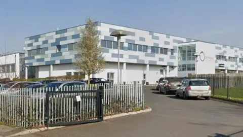 Exterior view of a school building, The building has a flat roof, with blocks of grey, blue and white on the walls. There is a metal fence in the foreground.