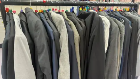 Annabel Amos/BBC A row of suits on a rail in different colours from grey, black and blue. They all have different size number tags on them. 
